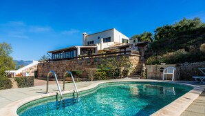 Una piscina con un muro de piedra, una terraza de madera y una casa con fachada de piedra y un balcón.