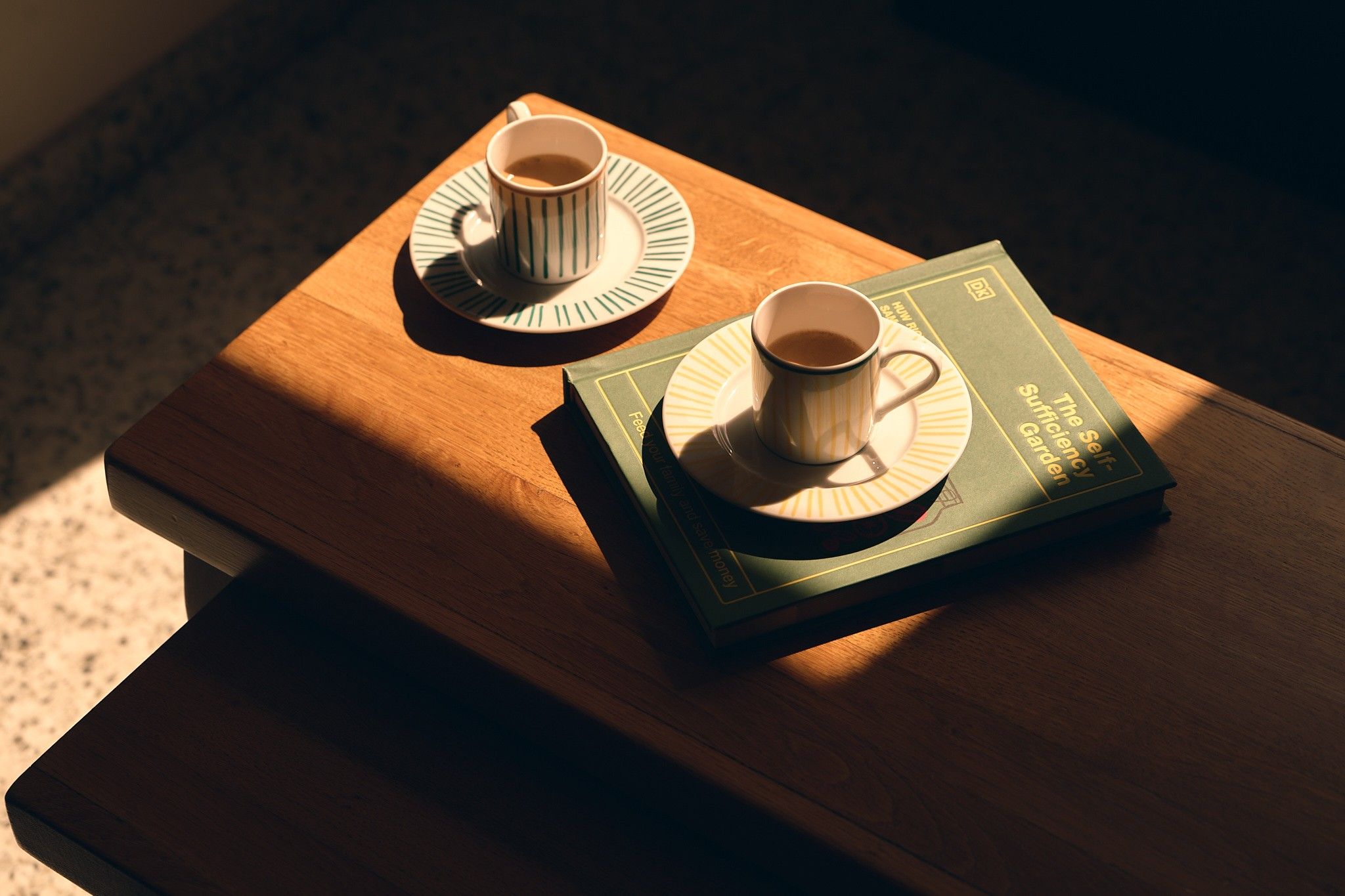 Un tavolo di legno con due tazze di caffè sui piattini e un libro intitolato 'The Sufficient Self-Garden'.