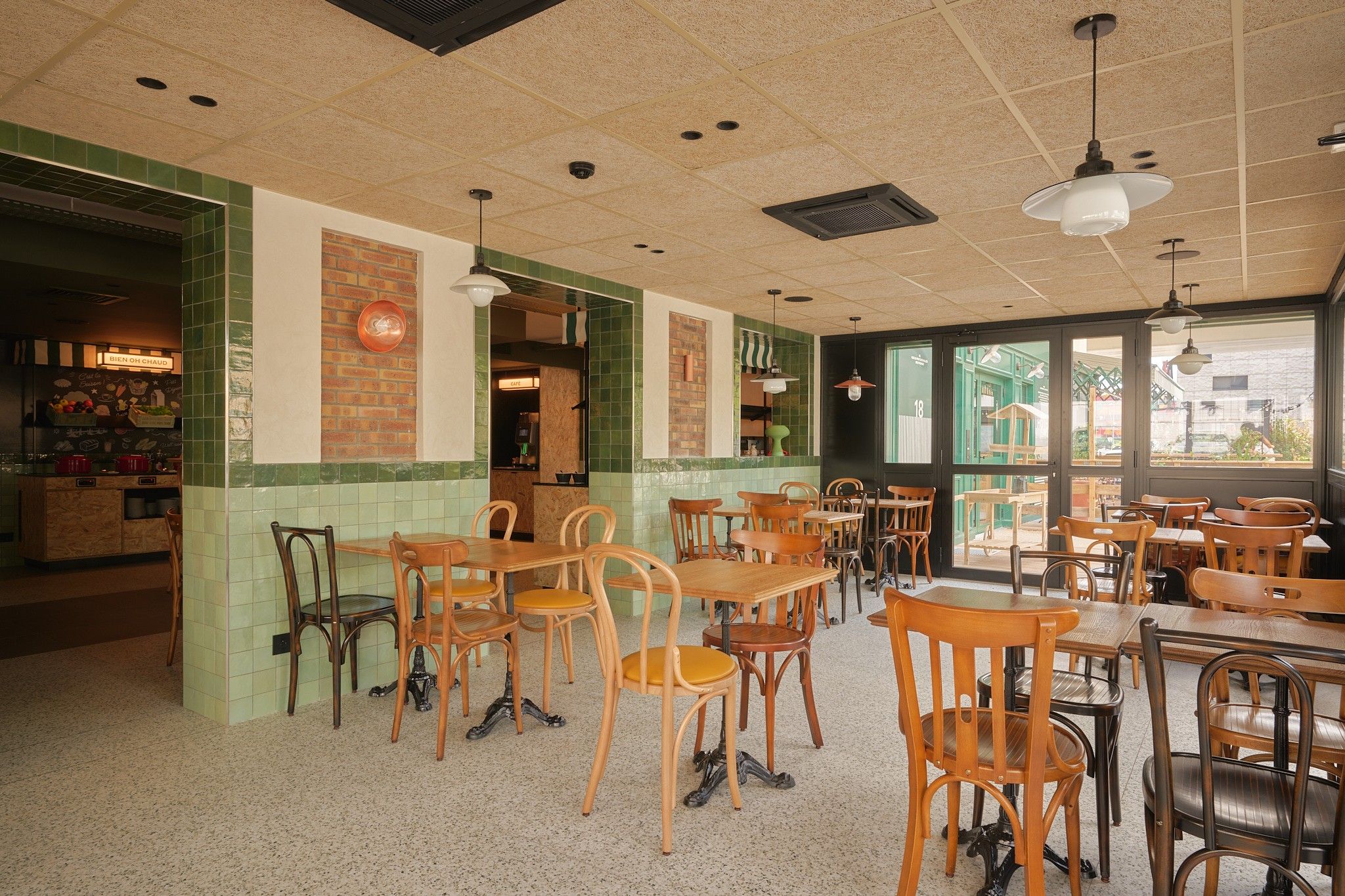 Een restaurantinterieur met houten tafels en stoelen, een wand betegeld met groene tegels en een uitzicht door grote ramen.