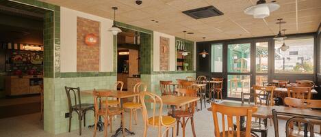 A restaurant interior with wooden tables and chairs, a green-tiled wall, and a view through large windows.