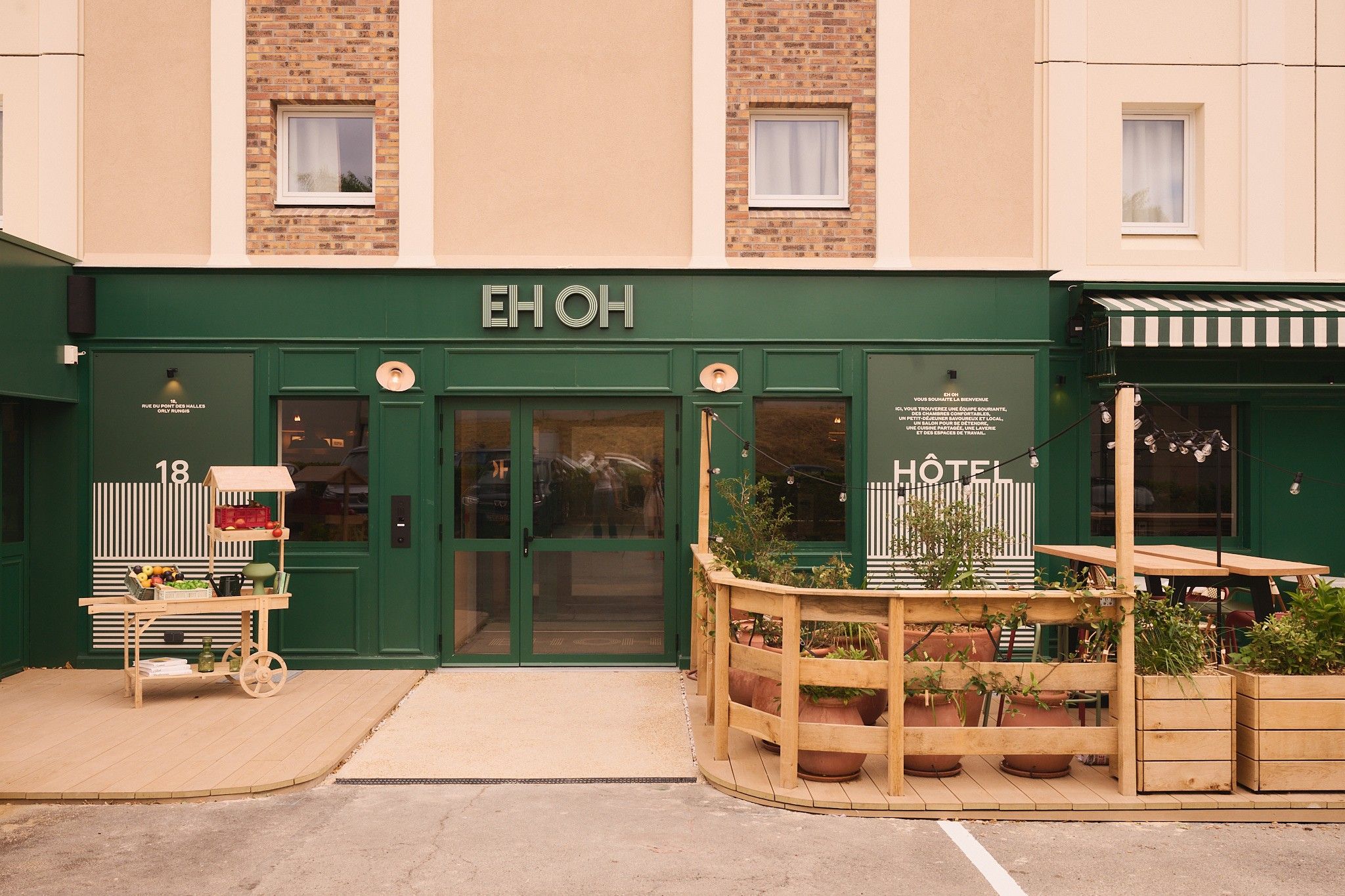 Entrada de un hotel con fachada verde, un letrero que dice 'EH OH' y un pequeño espacio al aire libre con plantas en macetas.