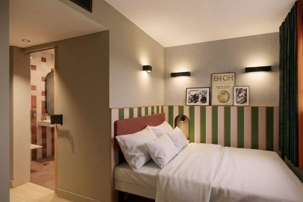 A hotel room with a bed, a wall-mounted light, a window with curtains, and a door leading to a bathroom.