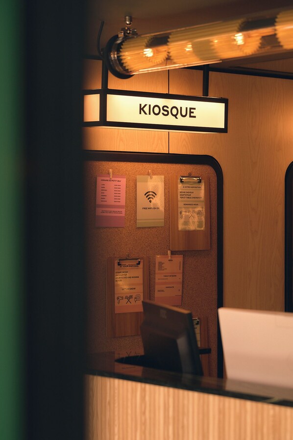 A reception desk with a sign reading 'KIOSQUE' and various posted notices including Wi-Fi availability and amenities like a hair dryer and ironing board.