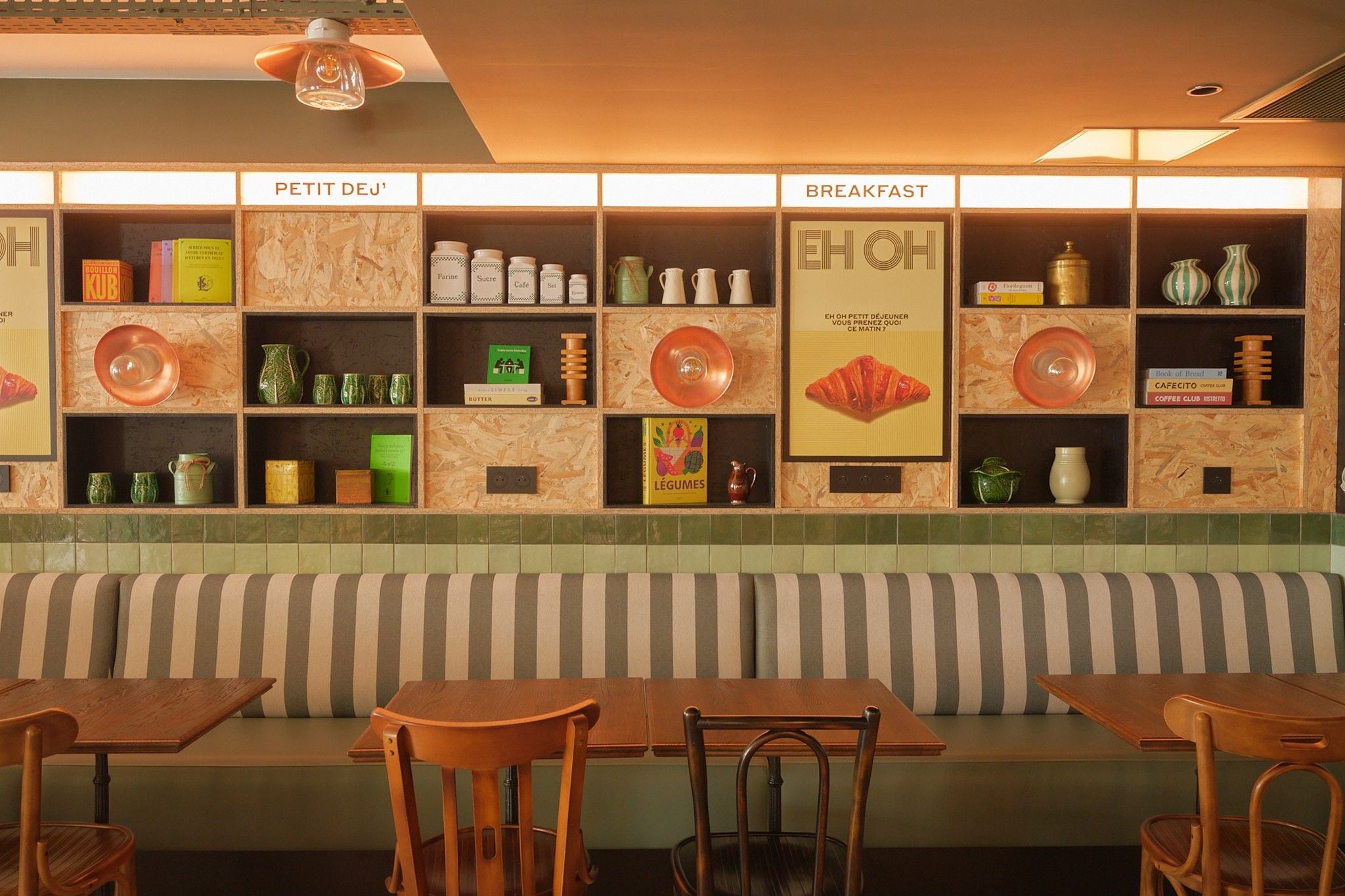 A restaurant interior with a wooden bench seating area, shelves with food items, and a wall with labels indicating sections like 'PETIT DEJ''.