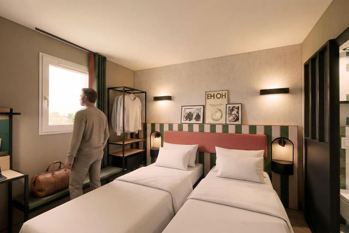 A hotel room with two beds, a desk with a laptop, a hanging coat rack, and artwork on the wall.