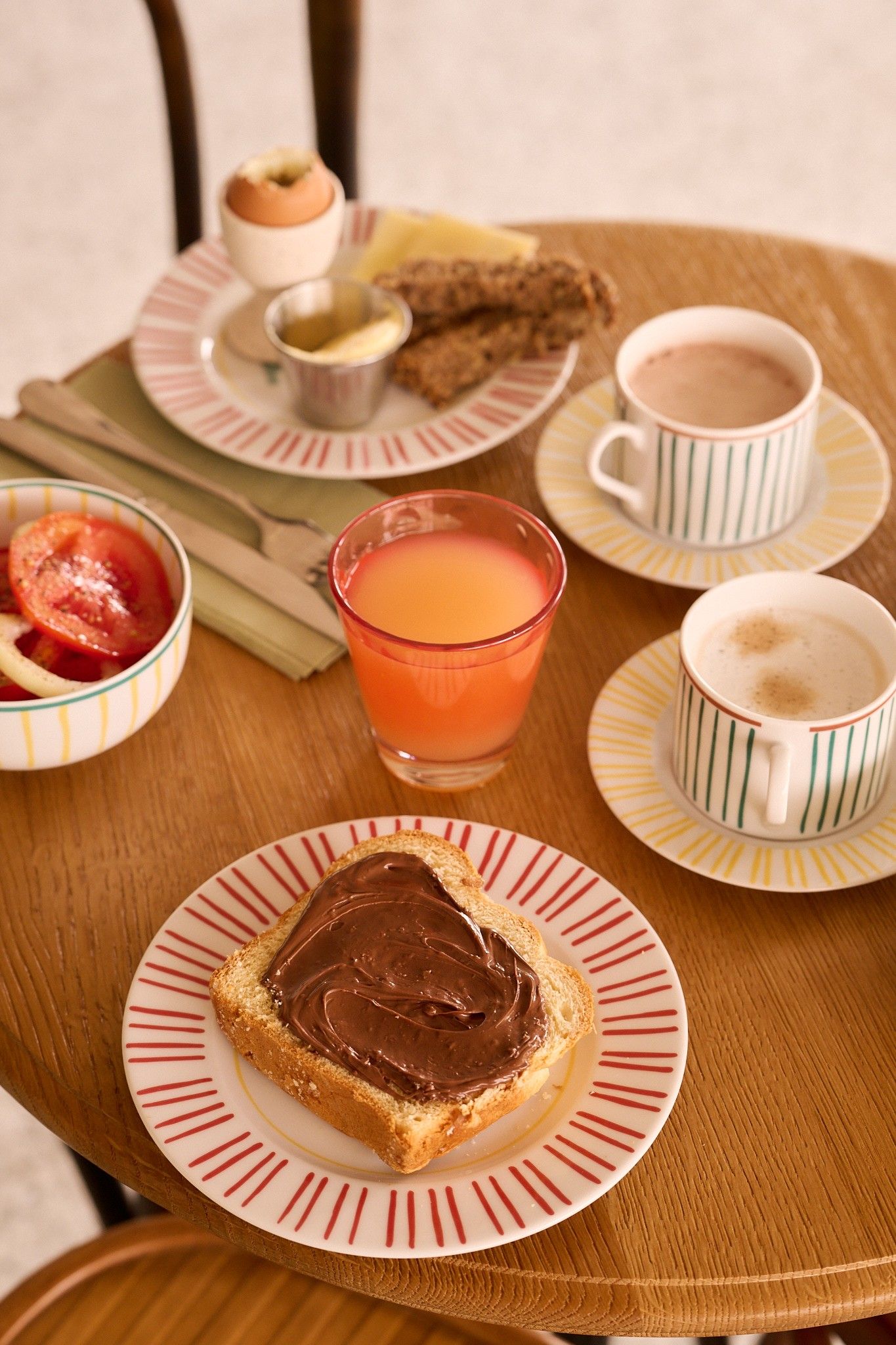 A table with a plate of toast with chocolate spread, a glass of orange juice, two cups of coffee, a plate with a muffin and a small bowl of jam, and a plate with a sandwich.