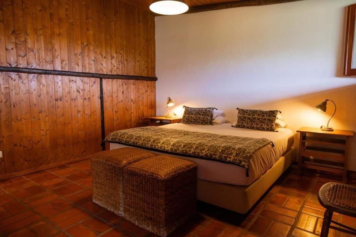 A bedroom with a wooden ceiling, a bed with patterned bedding, two bedside lamps, a wicker ottoman, and a chair.