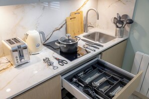 A kitchen with a marble countertop, a toaster, a kettle, a pot, a sink, a cutting board, and a drawer organizer with utensils. - 108 Shirland Road (London)