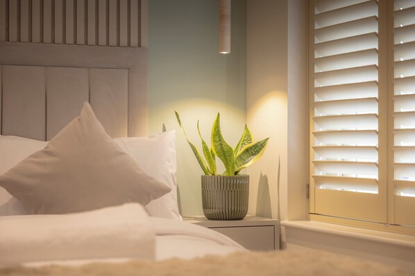 A bedroom with a bed, pillows, a bedside table with a potted plant, and a window with blinds. - 108 Shirland Road (London)