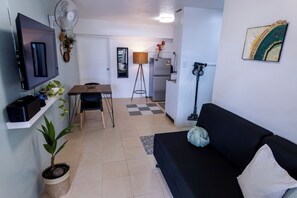 A compact living space with a TV, a dining table, a sofa, a refrigerator, a fan, and a potted plant.