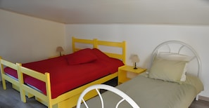 Ein Einzelbett mit roter Matratze und weißem Kopfteil, ein Nachttisch mit Lampe sowie ein Fenster mit Vorhängen.