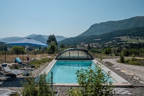 Una piscina con cerramiento de vidrio, rodeada de sillones de descanso y una sombrilla azul.