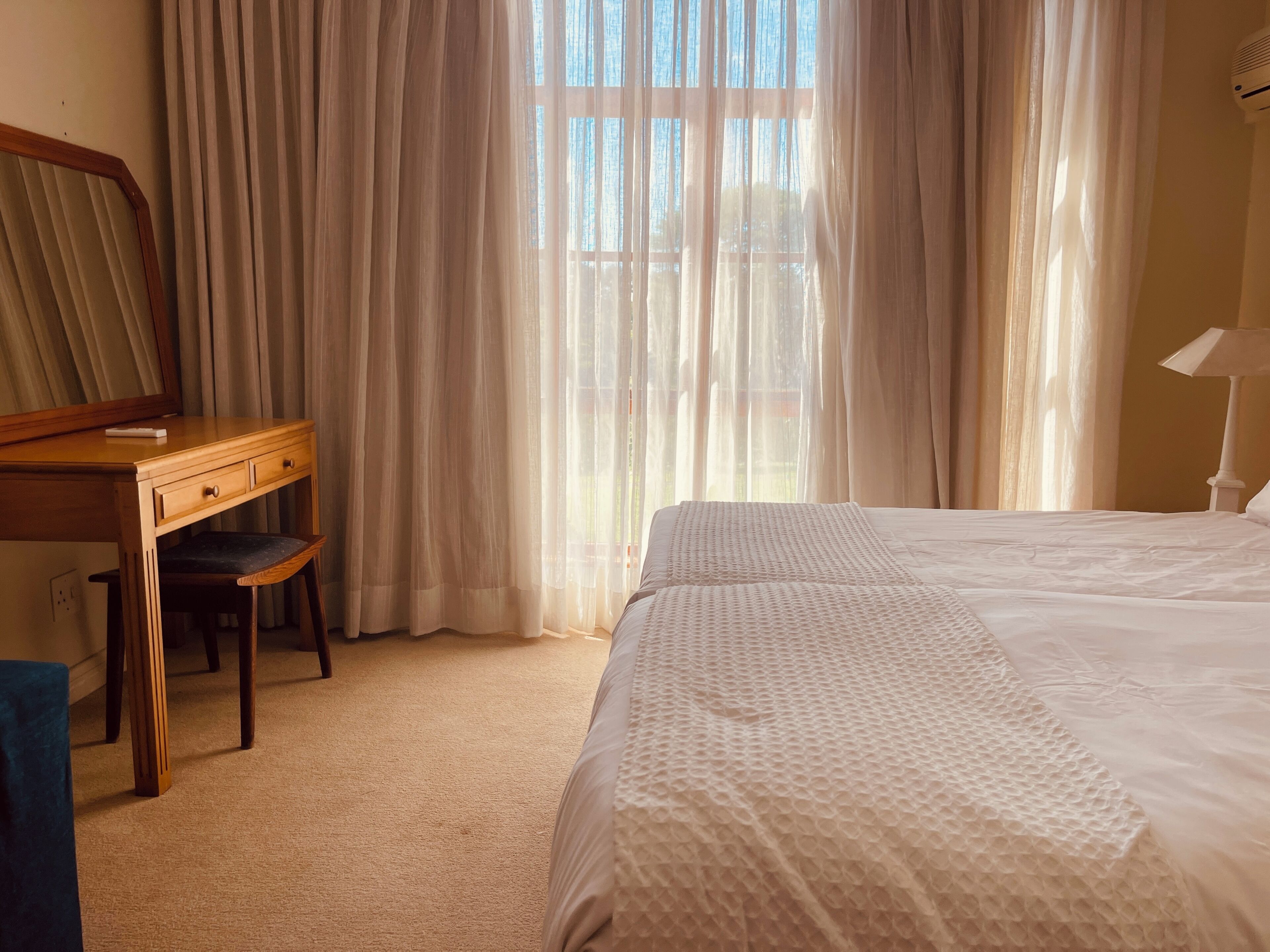 A hotel room with a bed, a desk with a chair, a mirror, and a lamp.