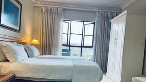 A neatly made bed with a headboard, a bedside table with a lamp, and a view of the outdoors through a window with curtains. - Selborne Golf Estate Villa P15 (Pennington)