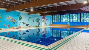 En inomhuspool med en väggmålning som föreställer marint liv, kristallklart vatten och utsikt över träd genom stora fönster.