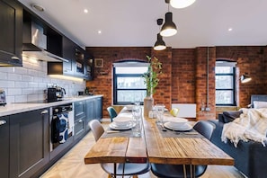A modern kitchen with dark cabinets, a wooden dining table set for four, and a brick wall backdrop. - Modern 2-Bed Flat in Central Leeds - Sleeps 6 (Leeds)