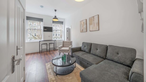 Uma sala de estar moderna com um sofá cinzento, uma mesa de centro de vidro, uma poltrona branca e vista para edifícios através de grandes janelas.