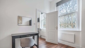 Um quarto com piso de madeira, uma escrivaninha com cadeira, uma janela grande com vista para edifícios e uma porta que dá para outro quarto.