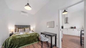 Um quarto com edredom de veludo verde, uma escrivaninha com cadeira e uma cozinha compacta no quarto vizinho.
