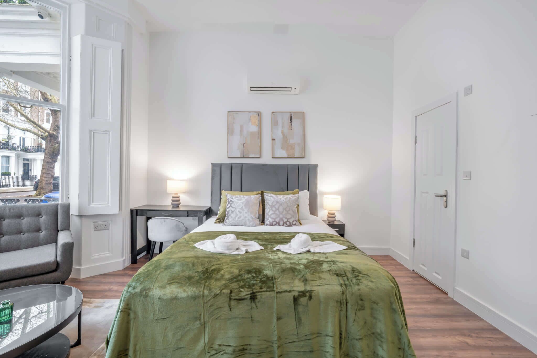 A hotel room with a bed, two bedside lamps, a desk with a chair, a sofa, and a view of the outside.