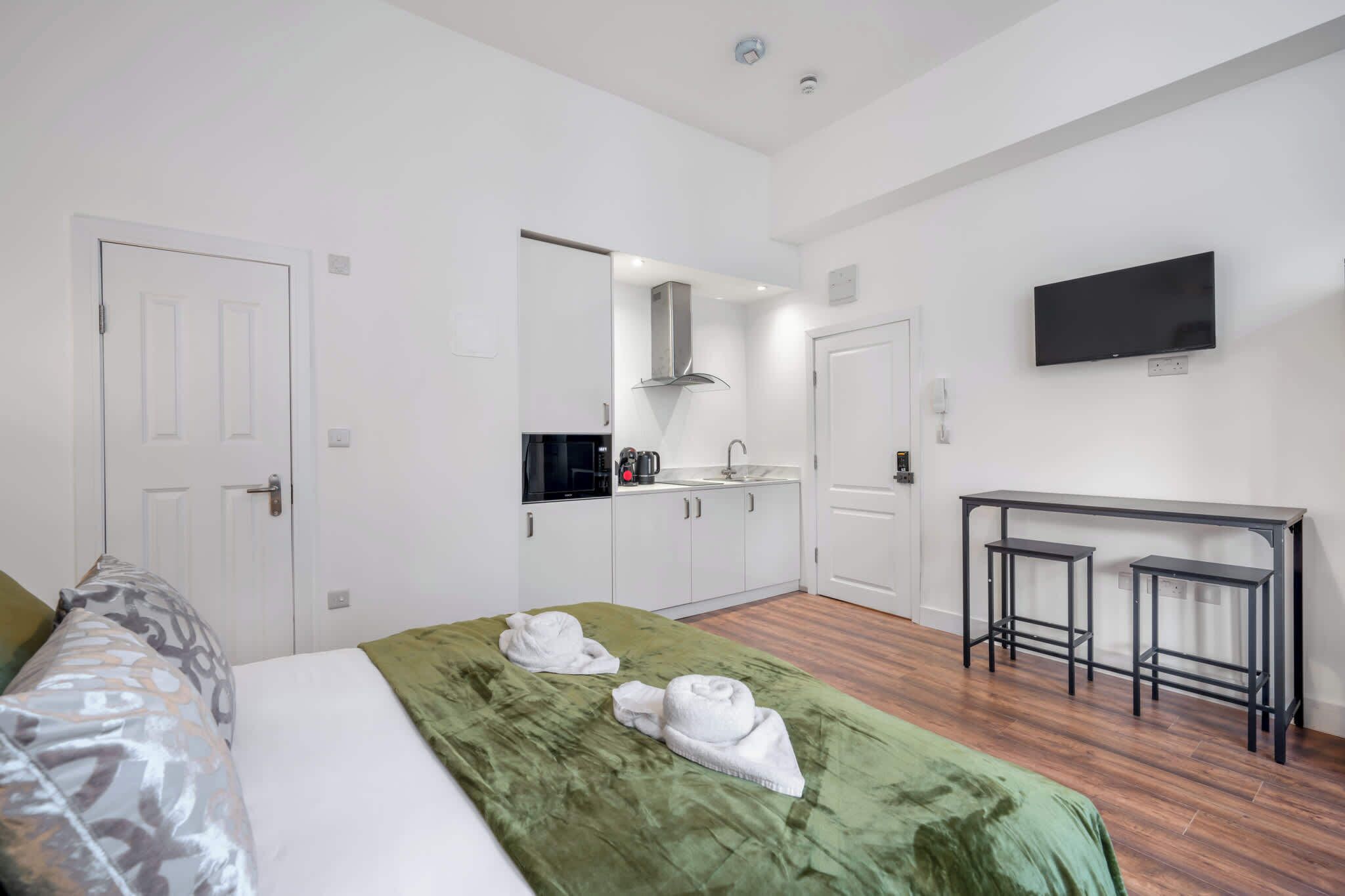 Une chambre d’hôtel avec un lit, une couvre-lit verte, deux serviettes, une kitchenette, une télévision et une petite table.