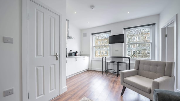 A modern kitchen with white cabinets, a dining area with a table and chairs, and a sofa in a living area with large windows. - Charming One Bedroom Flat in Knightsbridge (London)