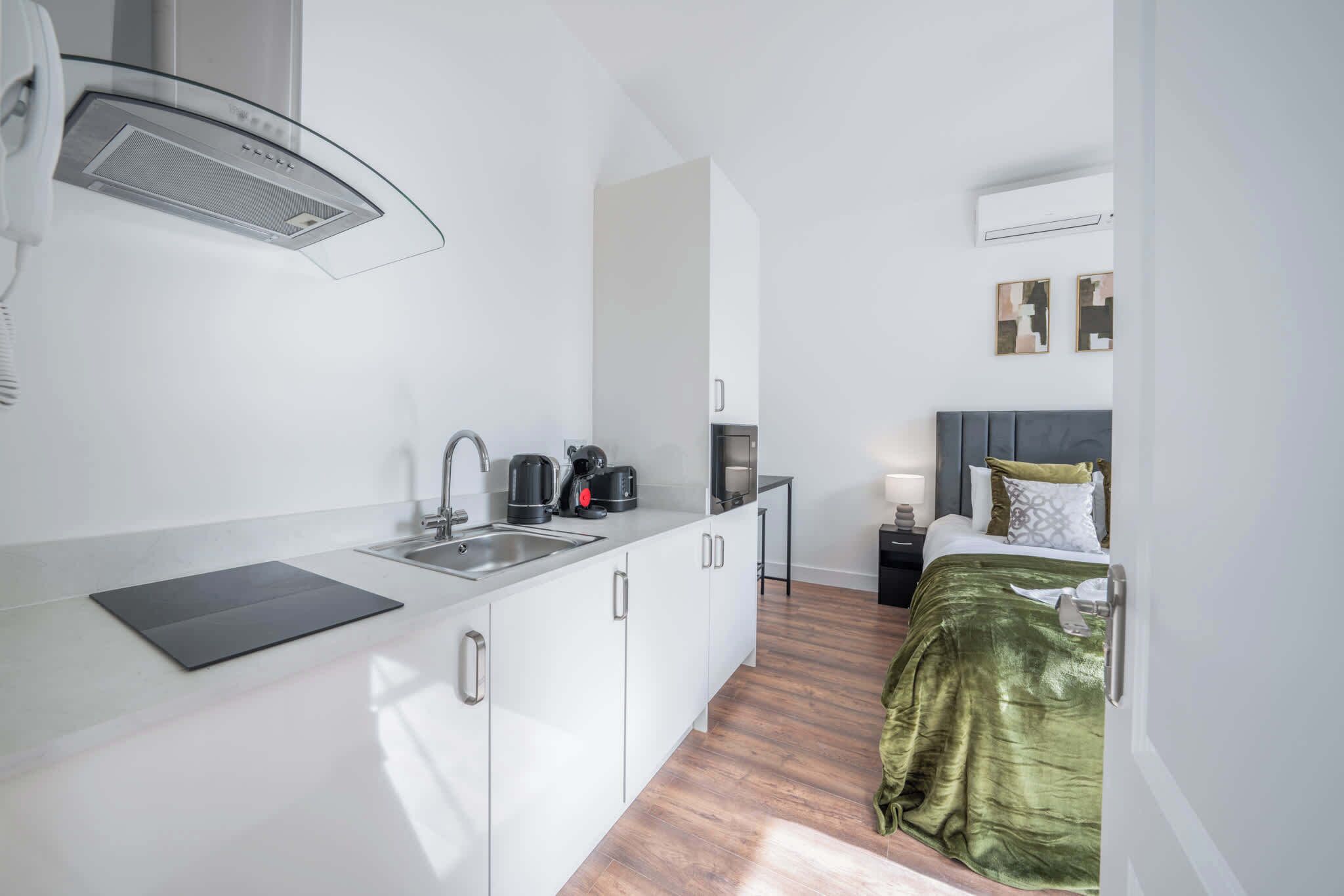 Een compacte keuken met een spoelbak, waterkoker en koffiezetapparaat; een bed met een groene deken; een nachtkastje met een lamp; en een wandairconditioner.