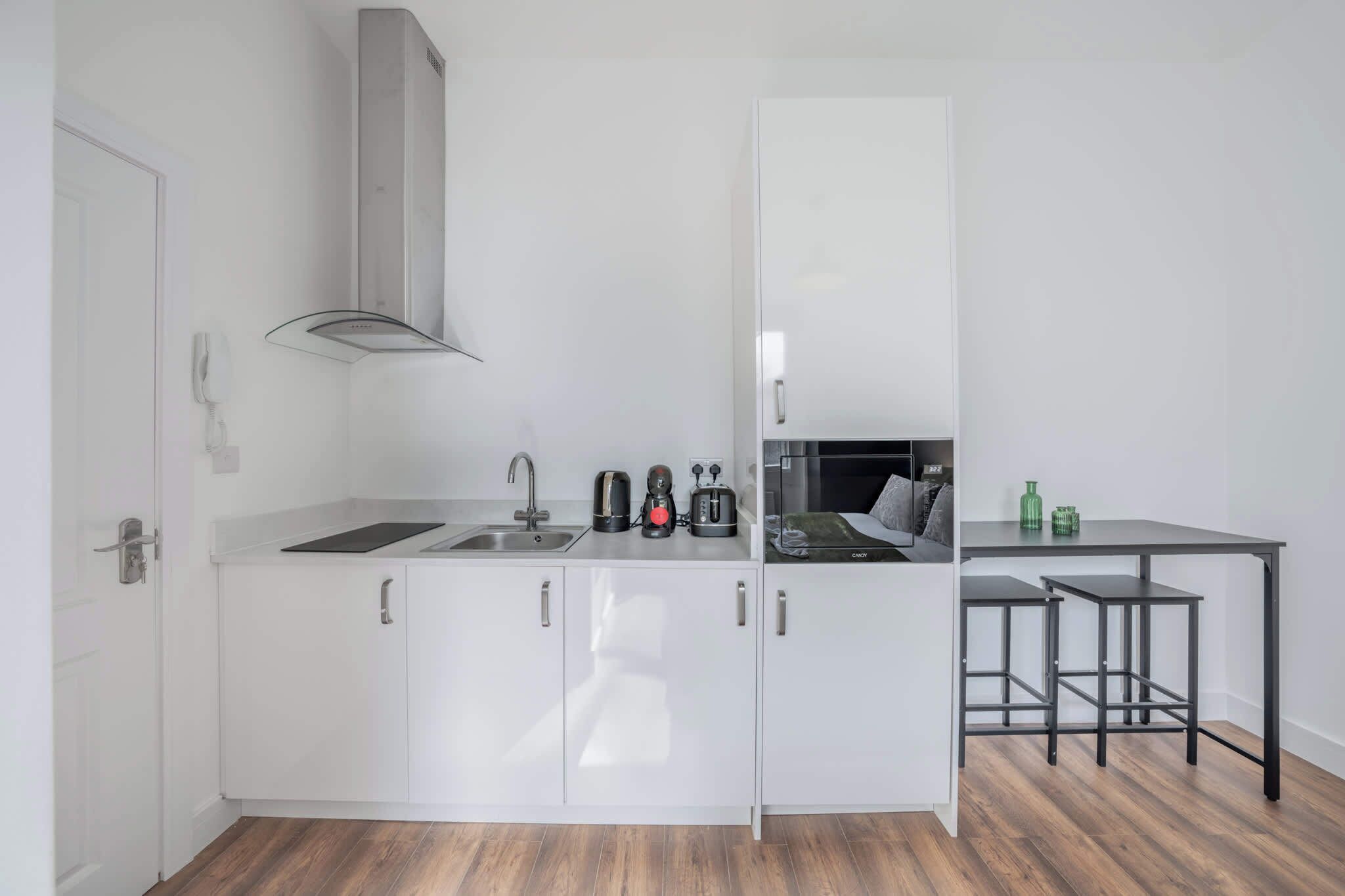 Une cuisine compacte avec des meubles blancs, un évier en acier inoxydable, une cuisinière à gaz et un petit coin repas équipé de tabourets.