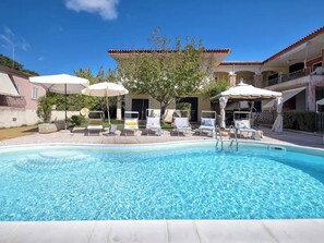 Una piscina con sillas de descanso y sombrillas, rodeada de edificios con tejados de tejas.