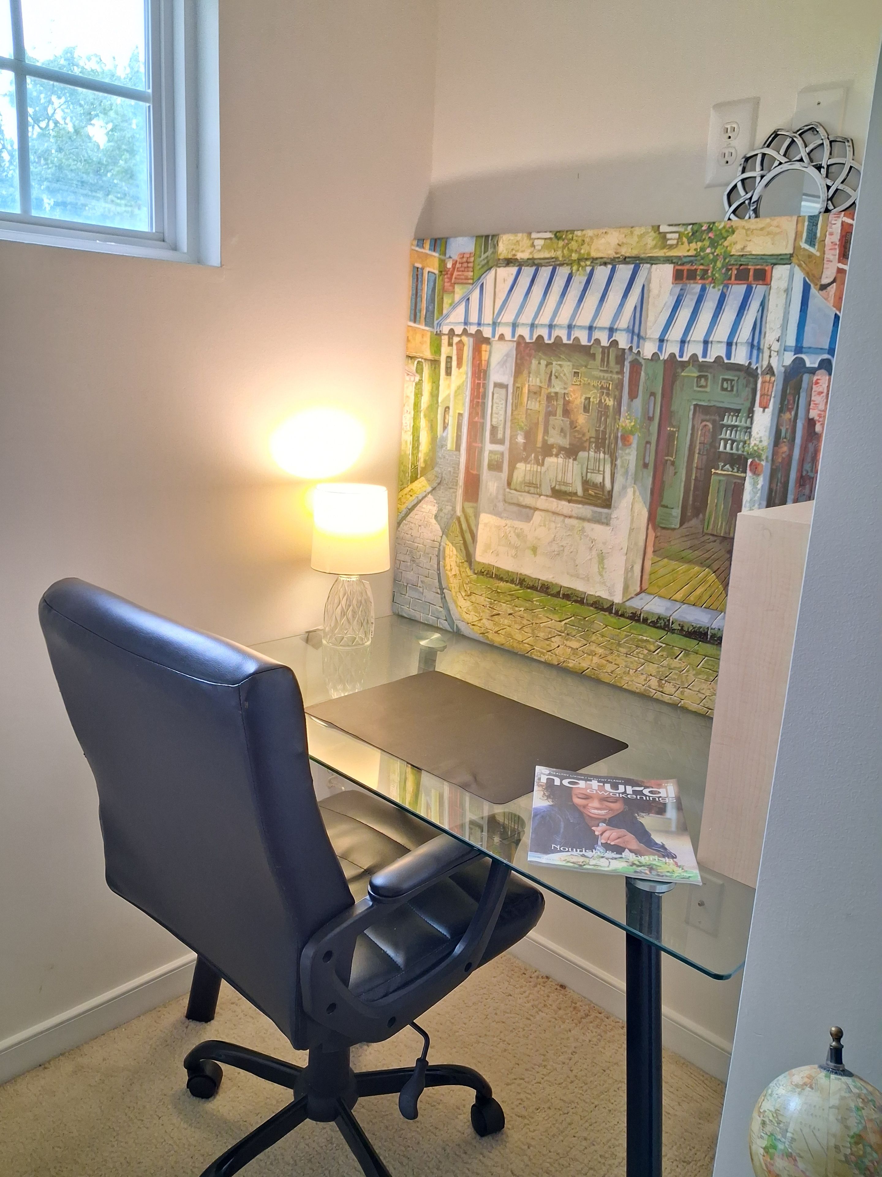 A room with a glass desk, a black office chair, a lamp, a wall mural, and a globe.