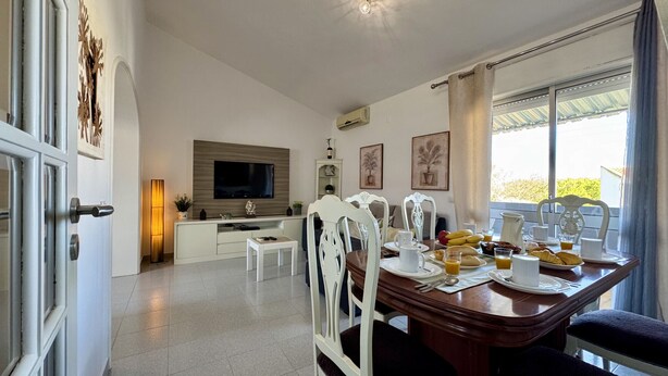 Un comedor con televisor, sillas blancas, una mesa de madera y alimentos para el desayuno.
