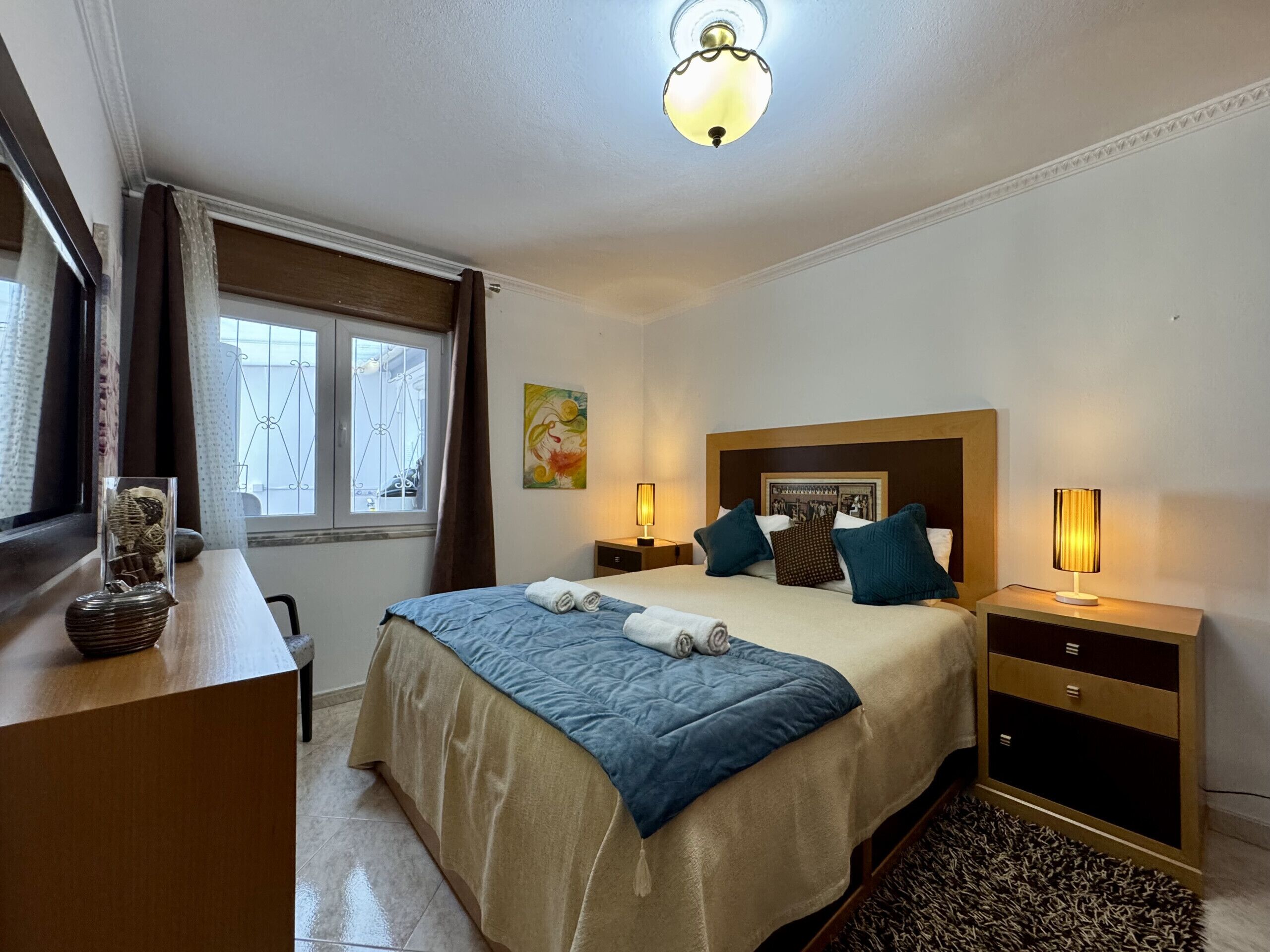 Une chambre d’hôtel comprenant un lit, des tables de chevet, un bureau, une fenêtre avec des rideaux, un luminaire au plafond et un tableau mural.