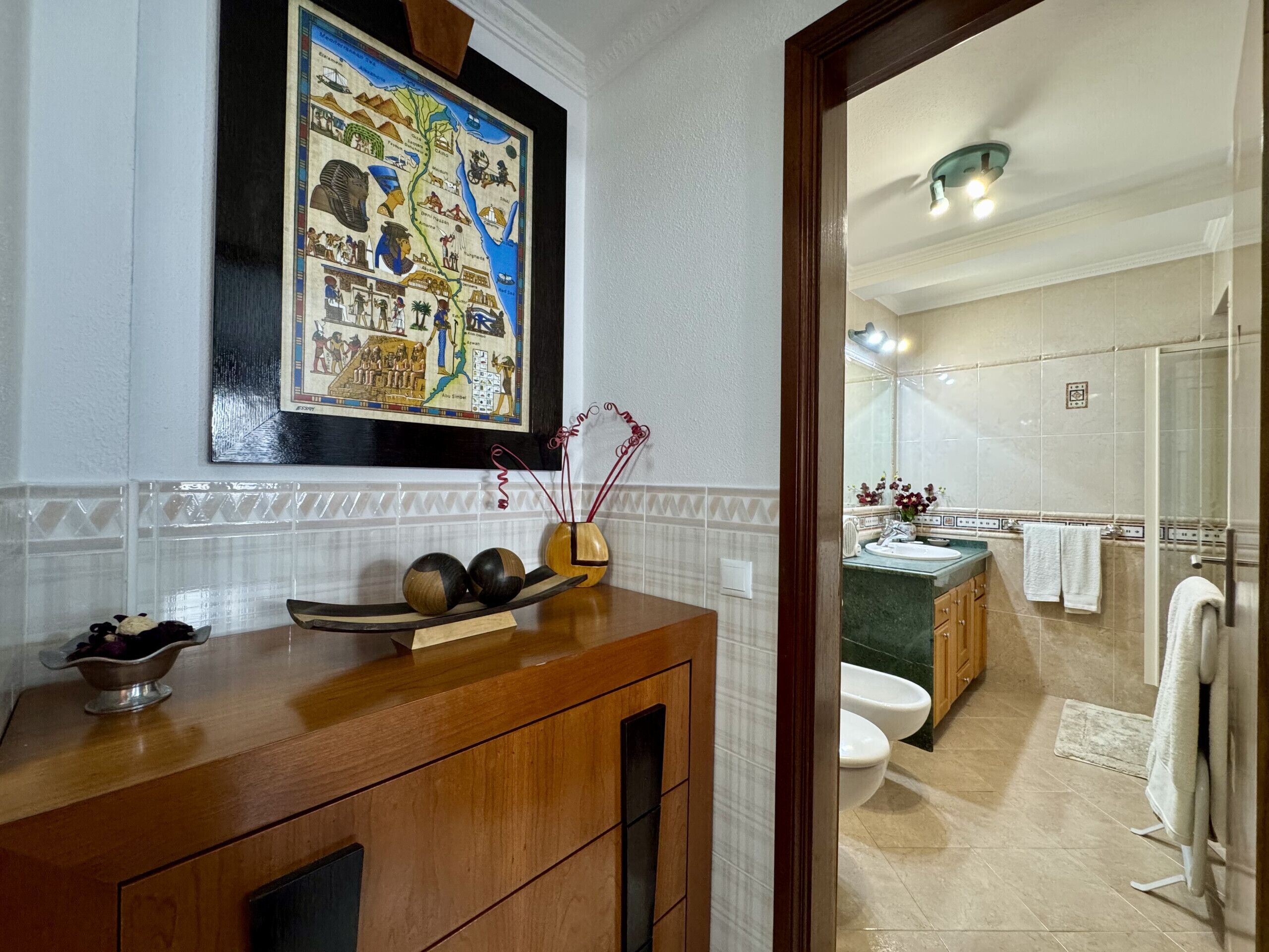 A bathroom with a wooden vanity, a decorative wall hanging, and a view into the adjacent room.