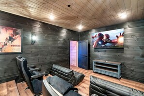 Ein Zimmer mit dunklen Holzwänden, Deckenbalken aus Holz und einem gro ßen Flachbildfernseher. Es bietet eine Sitzecke mit Ledersofas und einem Holztisch.