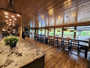 A spacious room with wooden floors, a long dining table with chairs, and large windows offering a view of the outdoors. - Luxury 15BR Cabin w/ Indoor Pool, Theater, Hot Tubs, Sleeps 64 Guests (Sevierville)