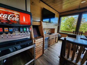 Un coin cuisine équipé d’un distributeur de Coca-Cola, d’un comptoir de bar en bois et d’une fenêtre donnant sur un espace verdoyant.