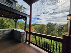 A covered outdoor deck with a stone fireplace, a wooden railing, and a view of a forested landscape. - Luxury 15BR Cabin w/ Indoor Pool, Theater, Hot Tubs, Sleeps 64 Guests (Sevierville)