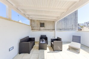 Een overdekt terras met rieten meubels en een kleine tafel, een betonnen muur en een heldere lucht.