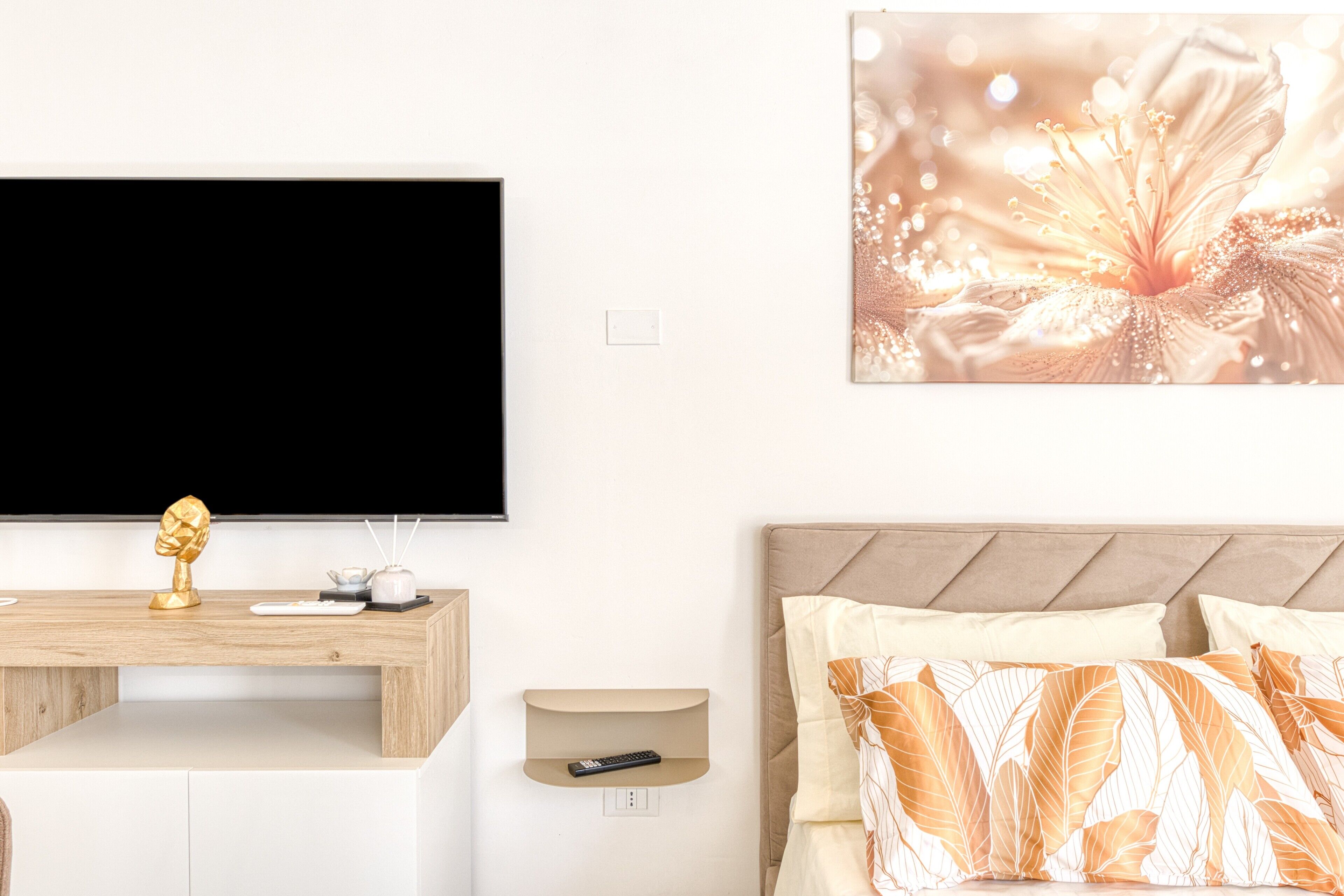Un dormitorio moderno con una televisión de pantalla plana, una mesita de madera, un estante para el control remoto, una cama con almohadas estampadas y una obra de arte en la pared.