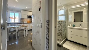 Eine moderne Küche mit weißen Schränken, ein Essbereich mit Tisch und Stühlen sowie ein Badezimmer mit Dusche und einem großen Spiegel.