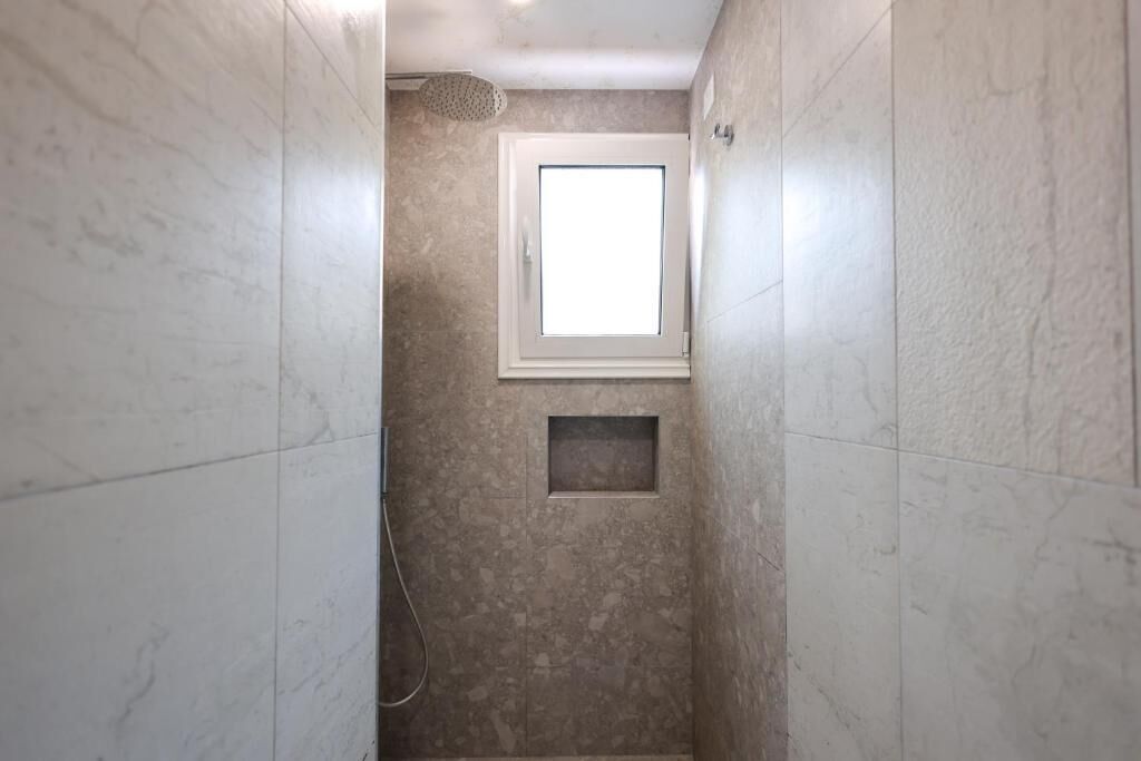 Een smalle badkamer met een douche, een klein raam en een inbouwplankje.