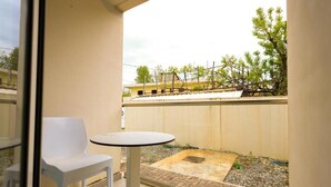 Un balcone con una sedia bianca e un tavolino rotondo, un pavimento in cemento e vista su un cortile recintato con alberi e un edificio sullo sfondo.
