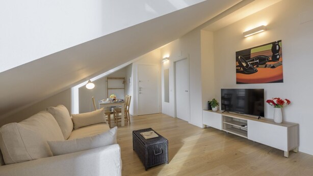 Un appartement moderne de style « loft », avec un plafond en pente, un canapé, un téléviseur posé sur un meuble blanc et un coin repas à l’arrière-plan.