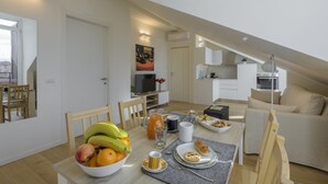 Une cuisine moderne avec des meubles blancs, une table à manger décorée de fruits, une machine à café et un téléviseur.