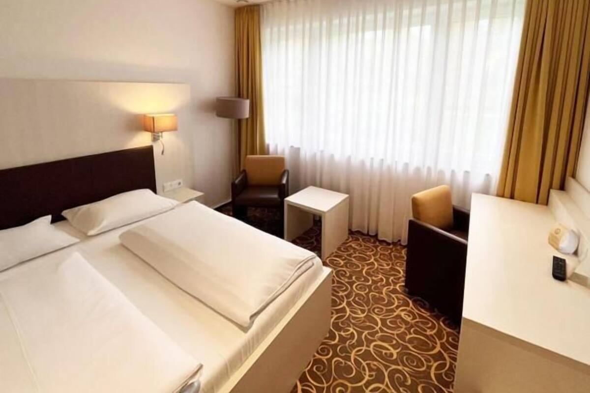 Une chambre d’hôtel avec un lit, un bureau, une chaise, une lampe et une fenêtre avec des rideaux.