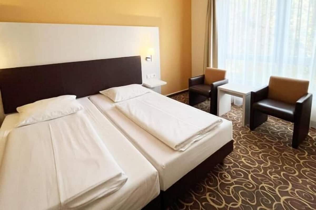 Une chambre d’hôtel avec deux lits, une petite table, une chaise et une grande fenêtre avec des rideaux.