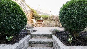 Un sentier de jardin mène à un espace aménagé avec des sièges, accessible par un escalier en pierre, et l’ensemble est entouré de haies bien entretenues et d’une clôture en bois.