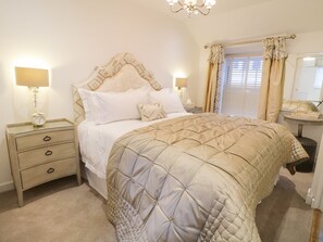 A bedroom with a bed, bedside tables, a nightstand, a chandelier, a window with curtains, and a mirror. - 8 Crown Lane (Conwy)