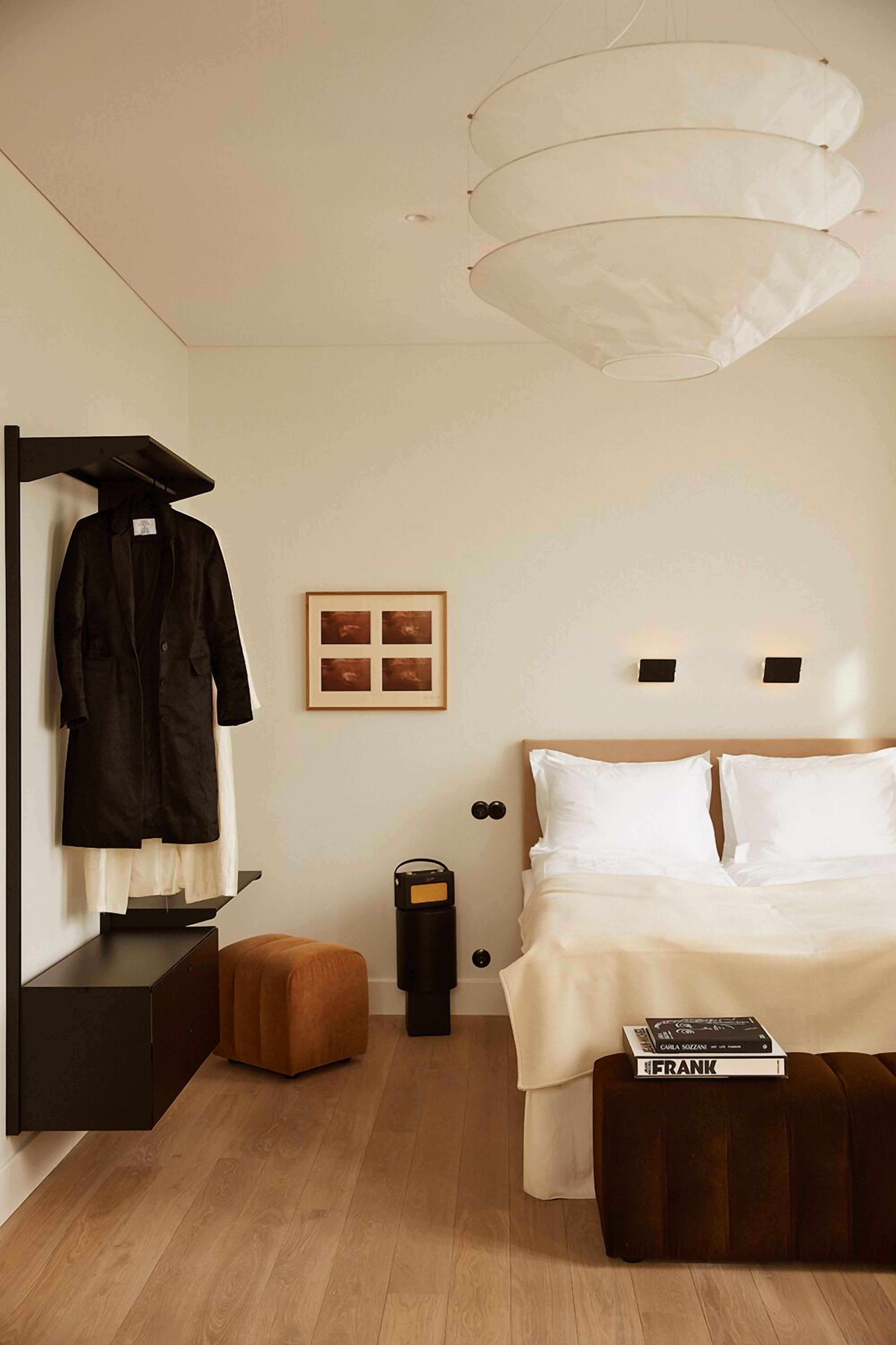 A minimalist hotel room with a bed, a coat rack, a framed picture, a small ottoman, and a hanging light fixture.