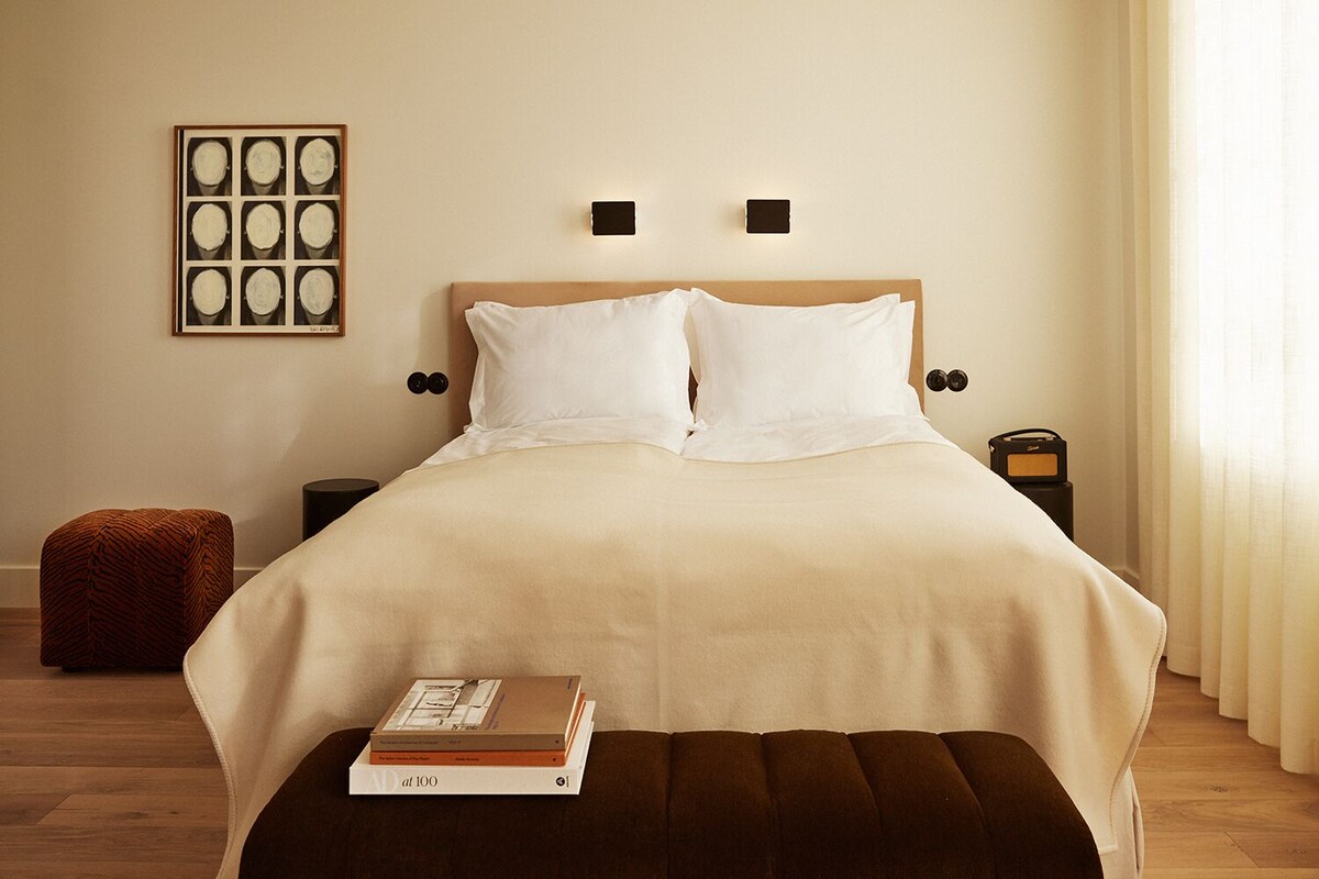 Un lit bien fait, recouvert d’une couvre-lit beige, avec deux oreillers, une table de chevet sur laquelle se trouve une radio, un petit pouf et un tableau encadré accroché au mur.